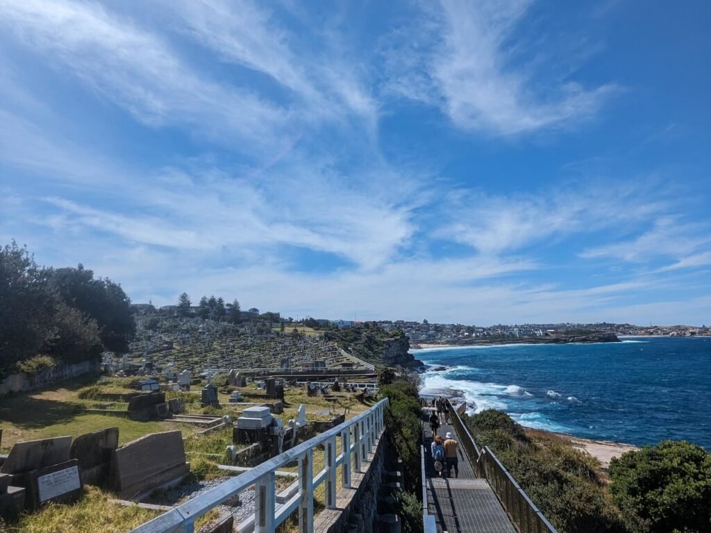 Walkers on a coastal boardwalk beside a cemetery, with the ocean to the right