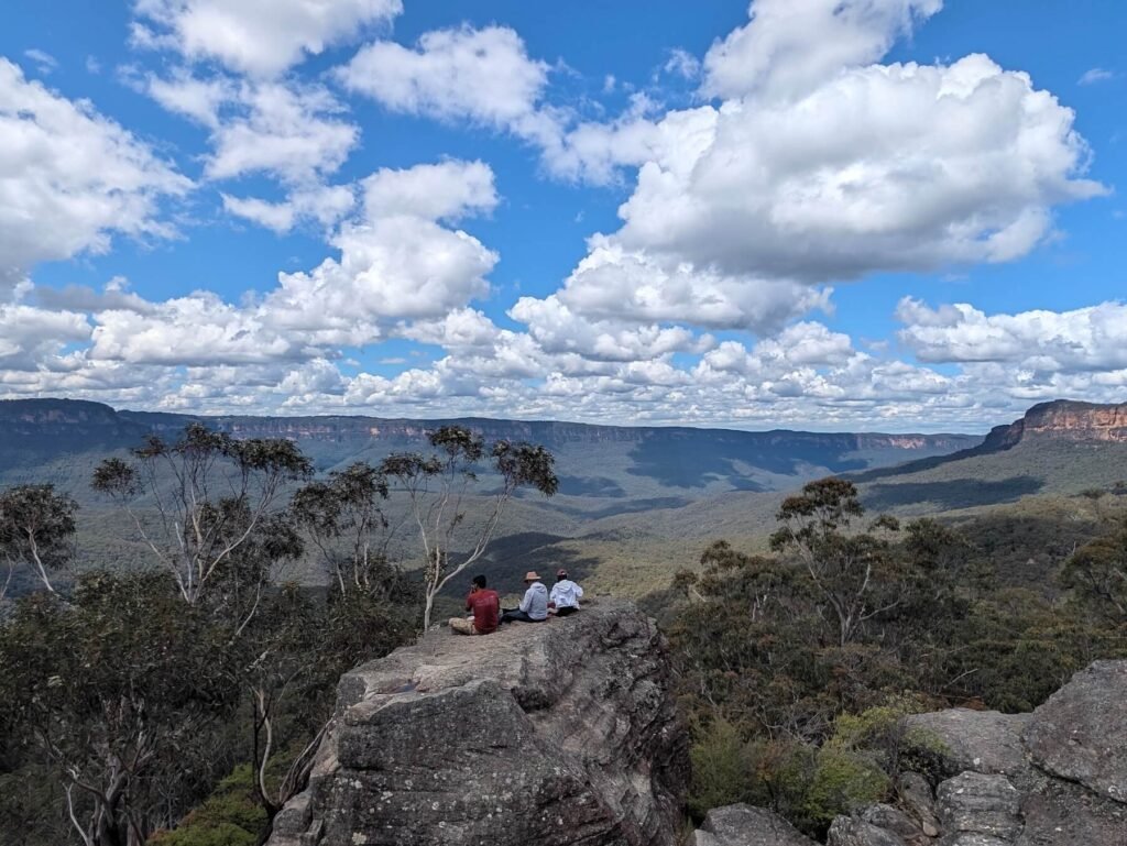 Walkers sitting on the rocks at Ruined Castle, looking across the Blue Mountains under a sky of white clouds